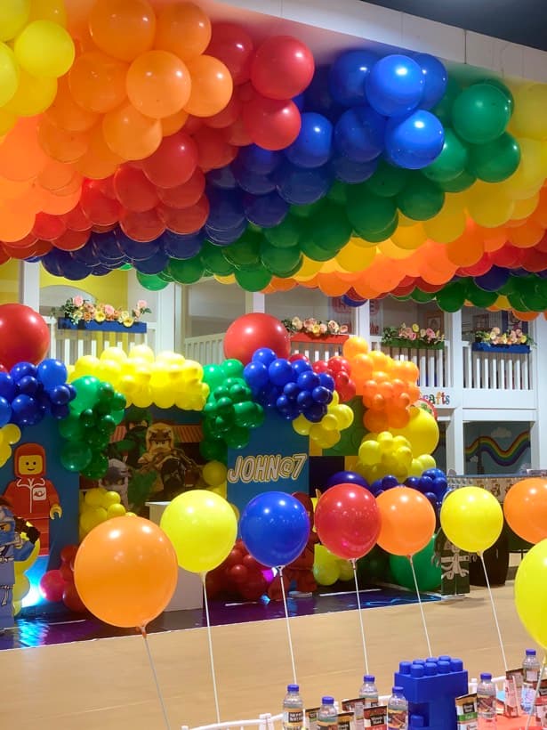 A Lego party setup with a rainbow balloon ceiling, brick backdrop and minifigure cut-outs
